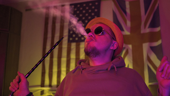 Young hookah smoker exhales up cloud of smoke in dark room with unusual neon glowing. Stylish man rests at home with American and GB flag on background.