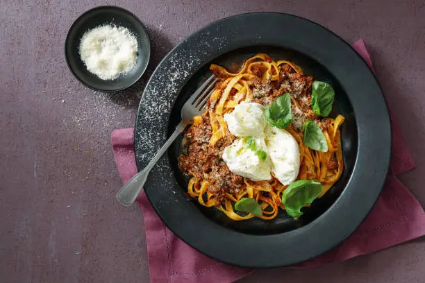 Rich Fettuccini Bolognese with Buffalo Mozzarella. Flat lay top-down composition on dark purple background. Horizontal image with copy space.