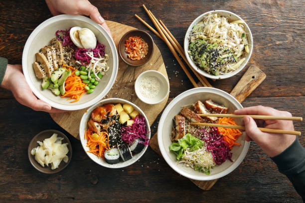 Family Eating Homemade Poke Bowls and Glass Noodle Salad Family eating homemade poke noodle salad with unagi eel. Poke bowl with fried tofu, vegetables, carrot, edamame, cabbage, wakame, mango and spicy shrimps on a bed of brown rice. Glass noodle salad with avocado and mango. Flat lay top-down composition on wooden background. junk food stock pictures, royalty-free photos & images