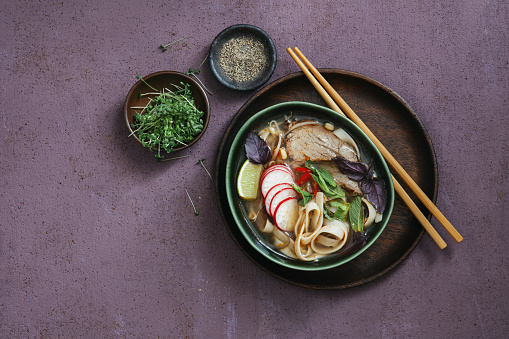 Pho is a Vietnamese noodle soup with rich broth, rice noodles, herbs, and beef. Flat lay top-down composition on purple background. Horizontal image with copy space.