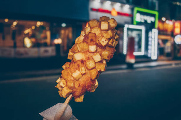 perro de maíz coreano hecho con patatas fritas con paisaje urbano nocturno borroso en el fondo - corn dog fotografías e imágenes de stock