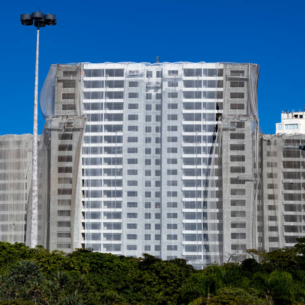 Urban construction site of large residential building draped in protective transparent veil with park vegetation in the foreground