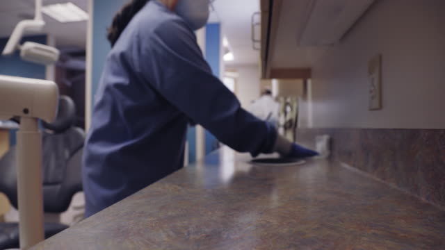 A Latina Dental Hygienist in Her Fifties Wearing a Face Mask Wipes off a Counter and Various Surfaces around an Examination Room at a Dental Office in Preparation for the Next Patient