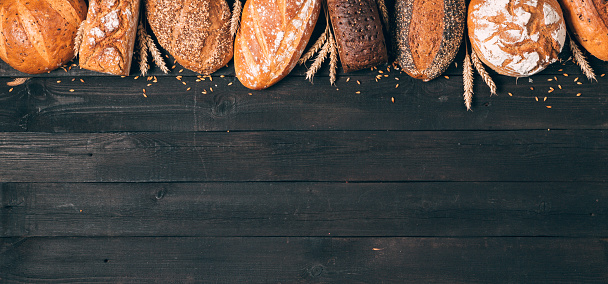 Bread on black wood background with copy space. Composition of black, brown and white whole grain loaves. Different types of bread from above. Panorama, banner. Bakery and grocery store concept.