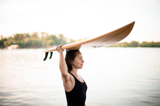 young athletic woman carries surfboard on her head. River and green trees in the background.