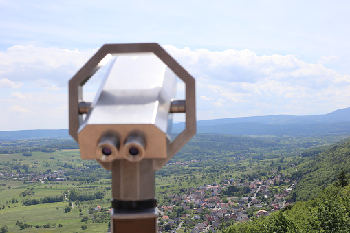 Stationary metal binocular against the backdrop of the mountains landscape and a green village in the valley. Telescope