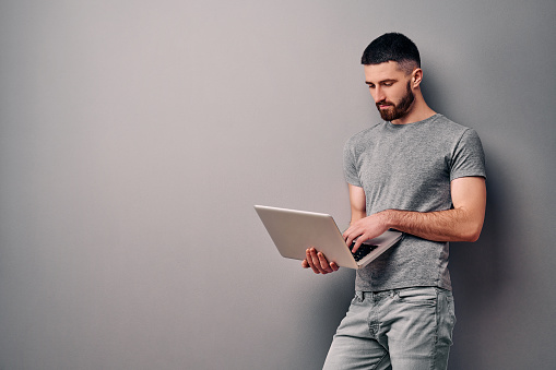 Portrait of young stylish handsome man in gray t shirt standing against gray wall with copy space for ads, holding laptop.