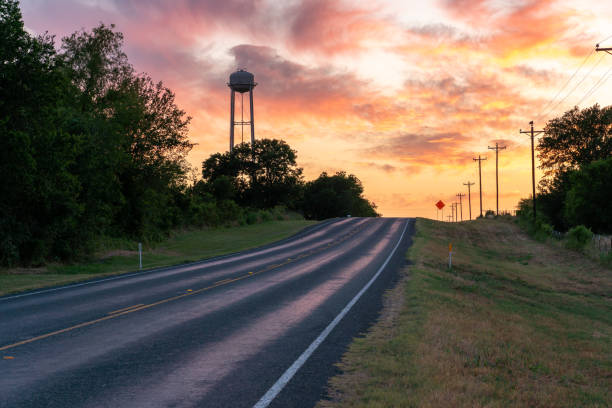 up hill road vers l’horizon avec tour d’eau au-dessus des arbres dans un coucher de soleil coloré - petite ville photos et images de collection