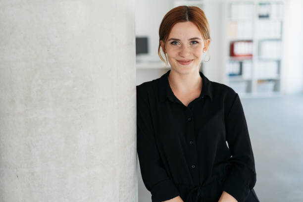 Friendly young businesswoman smiling at camera Friendly relaxed young businesswoman leaning against an interior office wall smiling at camera with lateral copy space employee introduction stock pictures, royalty-free photos & images