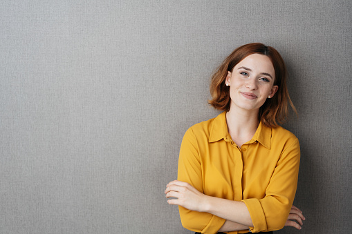 Pretty young woman grinning at the camera as she poses with folded arms against a grey studio background with copy space