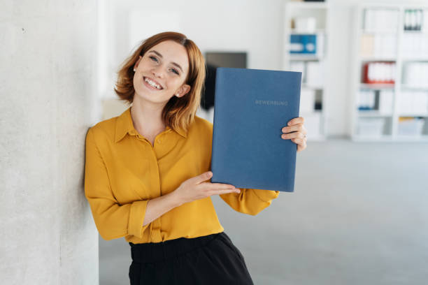 Young university graduate applying for a job Pretty young university graduate applying for a job holding up her CV in a blue file as she leans against an office wall smiling at the camera application form photos stock pictures, royalty-free photos & images