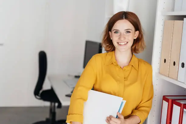Relaxed friendly young businesswoman holding a binder under her arm leaning against shelves in the office smiling at the camera