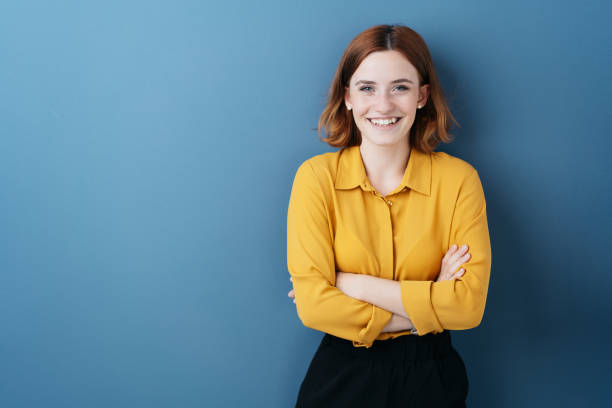 Pretty young woman with folded arms Pretty young woman with folded arms looking at the camera with a happy friendly smile against a blue studio background with copy space redhead photos stock pictures, royalty-free photos & images