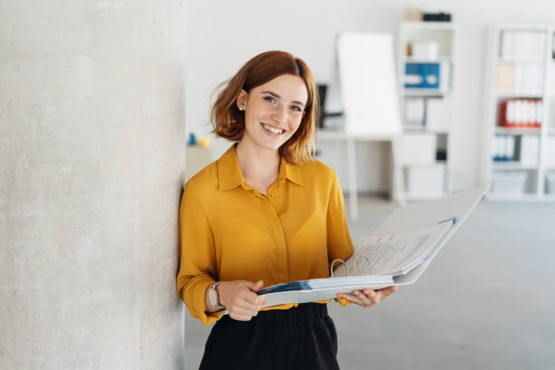 Attractive young office worker holding large file Attractive young office worker holding a large open binder as she looks at the camera with a sweet friendly smile redhead photos stock pictures, royalty-free photos & images