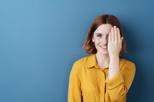 Attractive young redhead woman covering one eye with her hand as she peers at the camera with a happy smile against a blue studio background with copy space