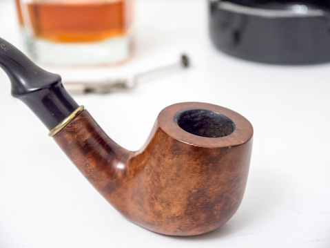 Briar brown smoking pipe with old fashioned glass of whisky, black ashtray and steel tool close side view with distorted background color image