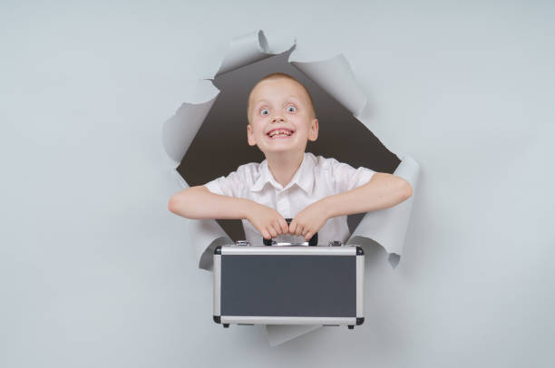 istock Cool boy is holding a suitcase full of money and emerging through torn paper hole in studio, has excited cheerful expression, looks through breakthrough of gray background. 1250936201