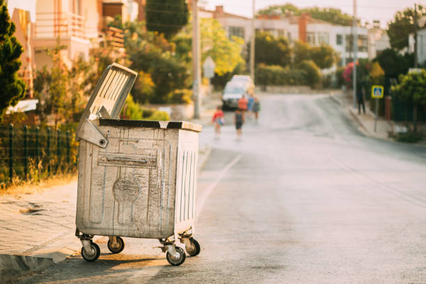 Metal Waste Container On Wheels In Street During Summer Sunny Evening. Container For Temporarily Storing Waste. Dustbin, Garbage Can, Trash Can