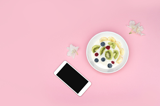 A smoothie bowl with superfoods and a phone on a pink background. Yogurt with blueberries, kiwi, banana and cereal. View from above.