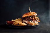 Juicy pulled pork burger on a plate with fries and onion rings. Dark background, copy space.