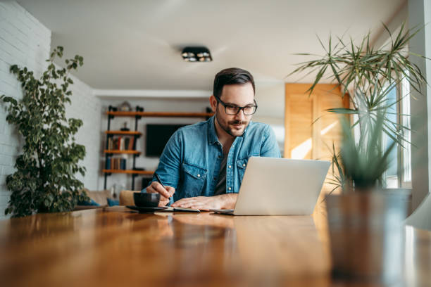 handsome man taking notes and looking at laptop, at home office, portrait. - house pen people caucasian imagens e fotografias de stock