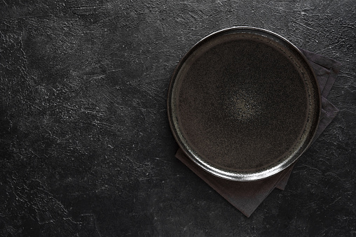Empty round black plate on dark black background, top view, copy space. Ceramic black platter on stone table, mockup.