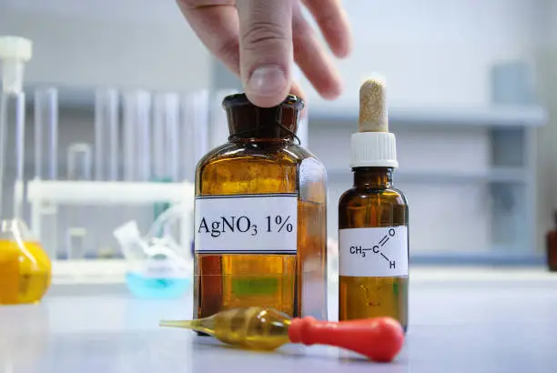 Photo of Scientist holding a bottle of silver nitrate with his fingers, flask of acetic aldehyde and glass dropper, ready for experiment, in chemical laboratory (blurred background)