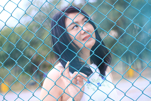 Beautiful asian woman in white dress sitting and smiling in natural park. Thai girl or Chinese Girl enjoy on holiday with sunlight in garden. Closed up focus on blue grating steel fence.