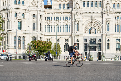 Madrid, Spain - June 15, 2020: Traffic in the Plaza de Cibeles in Madrid. A young man is riding a bicycle and they have a protective mask for the Covid-19.\