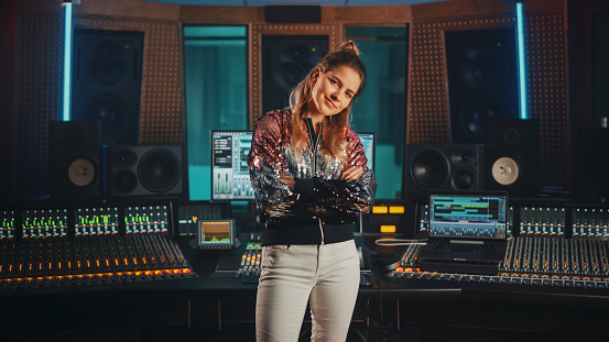 Portrait of Beautiful and Successful Caucasian Artist, Musician, Producer Wearing Stylish Jacket and Standing in Music Record Studio. In the Background Control Desk, Mixing Equipment, Soundproof Room