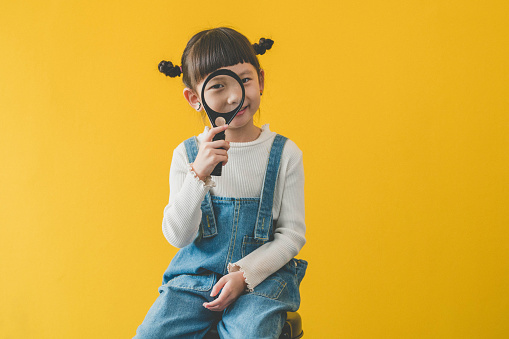 Cute Asian little girl holding magnifying glass smiling. A Little cute child girl looking magnifying glass on yellow background.