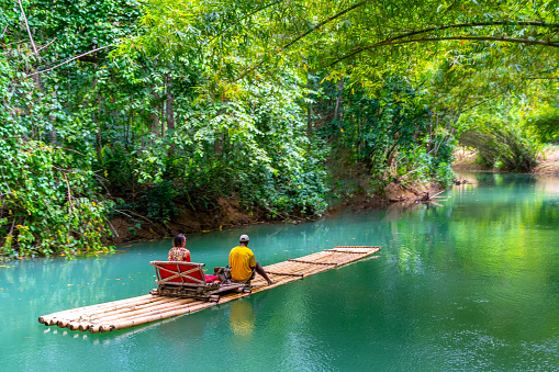 Falmouth, Jamaica - July 07 2019: Female tourist and tour guide sit on bamboo raft on Martha Brae River in Falmouth, Jamaica. Relaxing scenic ride through quiet countryside landscape under canopy of trees.