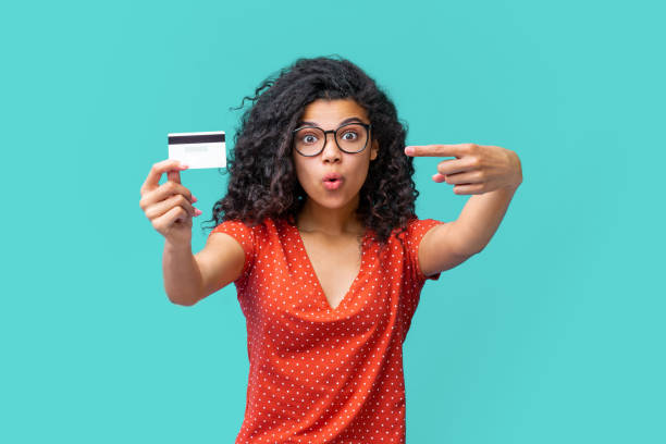 Studio portrait of happy attractive young african american woman in trendy spectacles smiling and holding credit card Cute surprised dark skinned girl in trendy spactacles pointing at credit card in her hand with index finger with wide-open eyes face expression. charging sports photos stock pictures, royalty-free photos & images