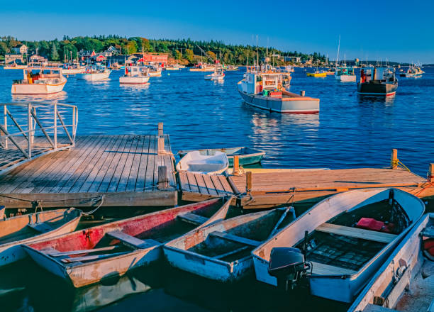 Fishing village Bass Harbor is warmly lit in the last part of the day. Row boats are tied to a wood wharf in front of fishing boats in New England's Bass Harbor, Maine. USA.  Last light of the day brings a glow to them all. fishing village stock pictures, royalty-free photos & images