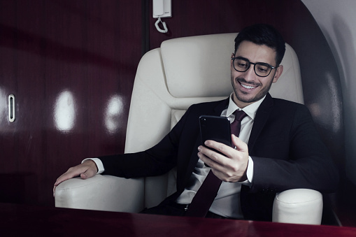 Young handsome businessman travelling first class, sitting in armchair next to window, reading text messages on smart phone screen and smiling happily