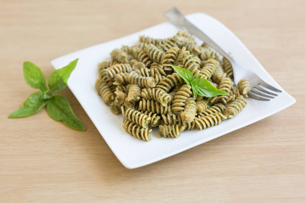 Top view of a white plate with spaghetti, pesto sauce and parmesan cheese surrounded by basil leaves, a silver fork and a napkin on a dark grey backdrop. The plate is at the center of the image. Low key DSLR photo taken with Canon EOS 6D Mark II and Canon EF 24-105 mm f/4L