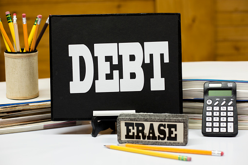 Word DEBT written on a chalkboard with an eraser near board. Pencils with erasers, books, bills, calculator featured in image.