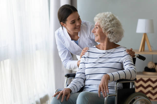 Smiling disabled elderly woman in wheelchair talking with caring nurse Smiling disabled elderly woman sitting in wheelchair talking with caring young nurse in living room, older generation receive homecare physical and moral support, caregiving and rehabilitation concept Engage Dementia Patients stock pictures, royalty-free photos & images