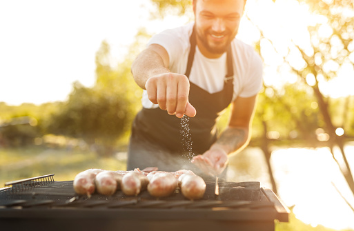 Crop male cook smiling and spilling salt of delicious fried sausages on grill on blurred background of countryside on sunny day
