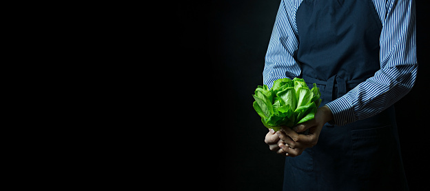 The cook in an apron holds a bunch of spinach in his hands. Healthy food. Vegetarian food. Dark background. Space for text.