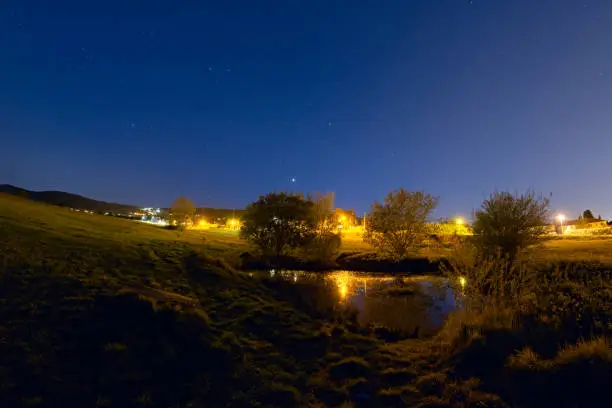 With the Malvern hills in the background, a lone bench looks out over a small pond with 4 trees that reflects the blue starry sky and streetlamps.