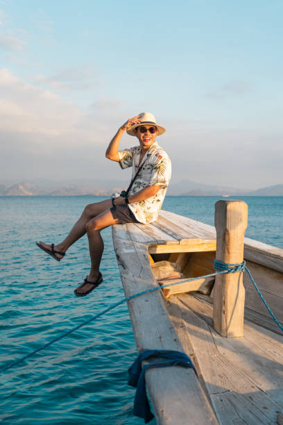 giovane viaggiatore felice seduto sulla punta della barca da pesca in un bellissimo tramonto serale. un uomo con la faccina sorridente. flores island in indonesia - flores man foto e immagini stock