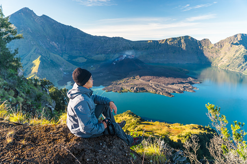 Young Asian man trekker sitting on Sembalun crater in Rinjani active volcano mountain, Lombok island in Indonesia, Asia