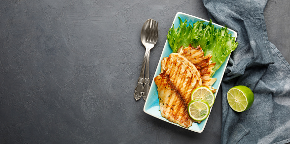 Grilled fish with fresh herbs and lime in a rectangular blue plate. Dark background. Top view with copy space.