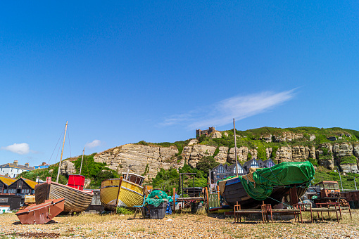 A view of Hastings Fishing Beach, one of the oldest shore launched fleets in the world. The view shows old boats, nets and gear strewn about. In the background Hastings Old Town, the East Hill and funicular railway can be seen, against a blue sky.