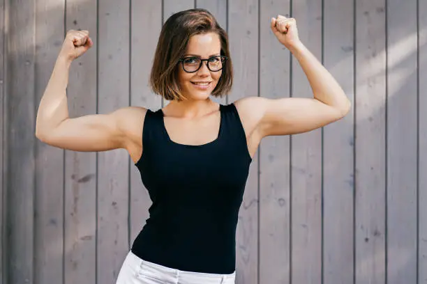Photo of Strong fitness girl with short hair dressed casual, demonstrates her biceps outdoor over wooden background
