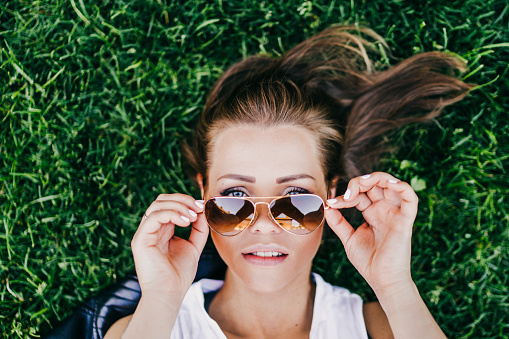 Close up portrait of beautiful young woman laying on green grass and looking over her aviator sunglasses holded by her hands. Summer time concept.