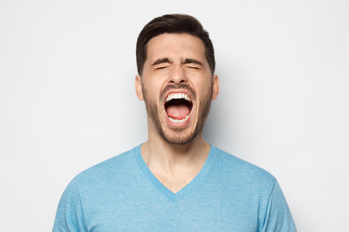Young man in blue t-shirt screaming loudly with closed eyes, feeling strong anger and despair, suffering from trouble and exhaustion, isolated on gray background