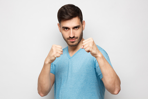 Smiling young man in casual blue t-shirt keeping his fists ready to boxing, isolated on gray background