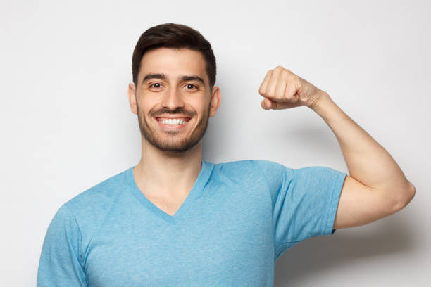 young strong sporty athletic man in casual blue t-shirt, showing biceps after training in gym, isolated on gray background - flexing muscles fotos imagens e fotografias de stock